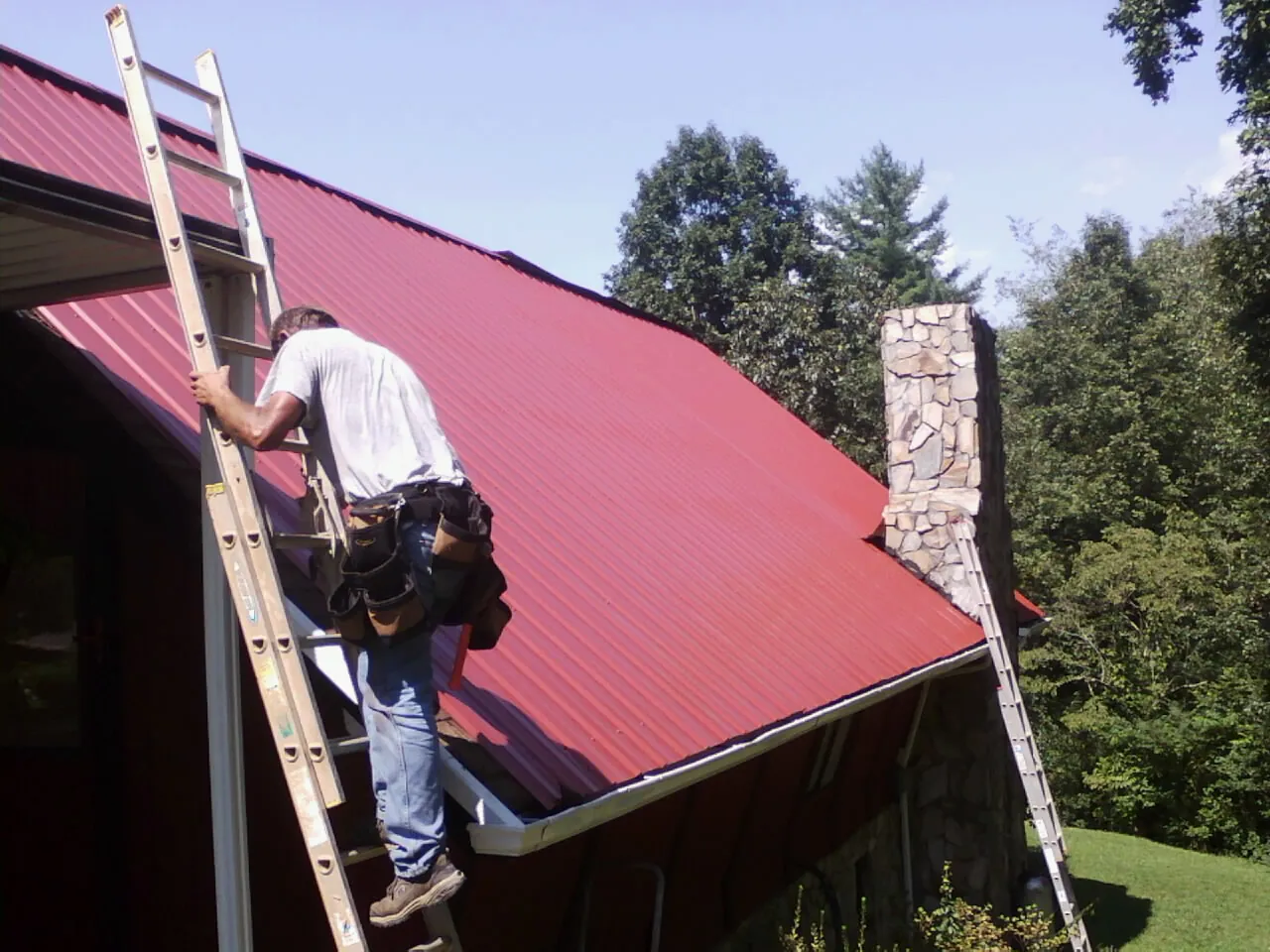Roofer on a ladder beside a red metal roof and stone chimney — Soffit & Fascia Repair by Roofing Brothers in Englishtown